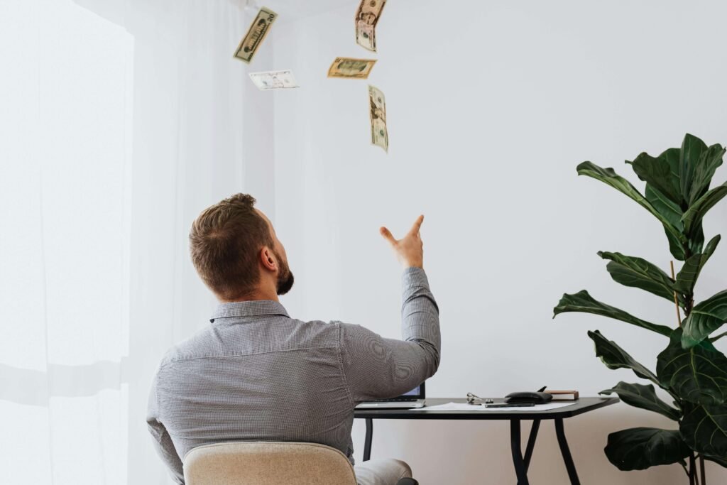 "A man in a light gray shirt tosses dollar bills into the air while seated at a modern desk, with a thriving fiddle-leaf fig plant—a popular and cheap Indoor Plants —adding vibrant greenery to the minimalist room."